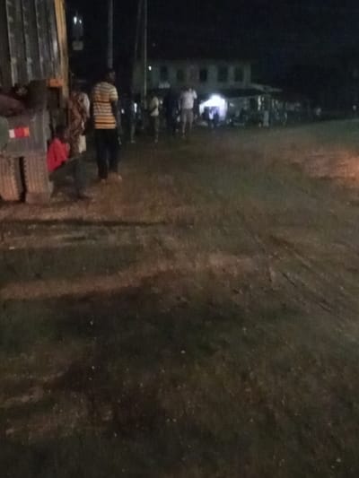 At night, a group of people gather near a dimly lit building. Some individuals stand close to a large truck, while others are further away, near the structure. The ground appears to be grass or dirt, showing signs of recent vehicle activity. The scene has a slightly blurry quality due to low light conditions.