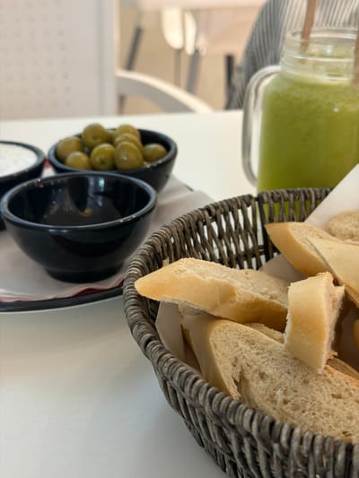 The image shows a close-up of a table setting, likely in a restaurant or cafe. The main objects in focus are a glass jar filled with a green smoothie or juice, a woven basket filled with sliced baguette bread, and two small black bowls containing green olives and possibly a dip or sauce.

The smoothie appears vibrant green and is in a clear glass jar with a wooden stirrer. The baguette slices are golden brown and appear fresh, resting in a dark woven basket lined with white paper. One black bowl is filled with plump green olives, while the other is empty, revealing a glossy black interior.

The background is blurred, but suggests an indoor setting with white tables and chairs. The lighting appears natural, hinting at daytime. The overall presentation suggests a light meal or appetizer service.

There is no visible text in the image. The scene is well-lit and the focus is sharp on the food items, indicating a deliberate composition for presentation. The context of Sant Antoni De Portmany, Spain, suggests a Mediterranean setting, which aligns with the olives and bread.