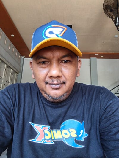 A close-up, outdoor, eye-level selfie shows a man wearing a black t-shirt and a blue and yellow baseball cap. The man has brown skin, a goatee, and is looking directly at the camera with a neutral expression. The t-shirt has a graphic print on it, appearing to be the character Sonic the Hedgehog. The background is out of focus but suggests an outdoor environment with a building and a fan mounted on a ceiling. The overall impression is that of a casual portrait.