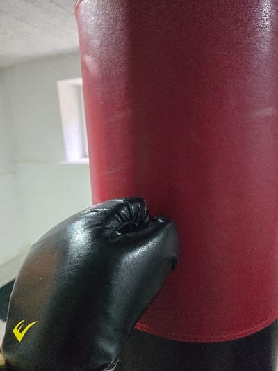 An indoor, close-up image displays a left hand encased in a black boxing glove, marked with a yellow stylized 'E' logo on the back, held in a clenched fist posture. The glove is positioned in the foreground, angled towards the right side of the frame. Directly behind the glove, and filling the right two-thirds of the image, is a large, cylindrical punching bag, predominantly red with a darker, possibly black, bottom section. In the blurred background, a light-colored wall, likely white, is visible, featuring a rectangular window aperture that admits ambient light. The setting appears to be a training environment such as a gym or workout space. No discernible features within the image indicate a specific location in the city of Kumanovo, North Macedonia.