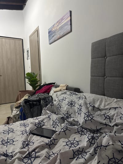 The image depicts a bedroom scene, likely in an apartment. In the foreground, a bed is covered with white sheets patterned with dark blue ship's wheels. A dark-colored smartphone lies on the sheets. Behind the bed, a large, upholstered grey headboard is visible. To the left of the bed, a sofa is covered with a pile of clothes and a potted plant with large green leaves. Two doors, one slightly ajar, are visible on the far left of the image, suggesting a hallway or entrance area. A framed picture of a seascape hangs on the wall above the sofa. The lighting suggests it is either dusk or a dimly lit interior. The location is identified as Asprovalta, Greece.