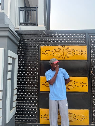 A man stands in front of an ornate gate. He is wearing a light blue shirt, gray pants, and a cap. He has one hand up to his face. The gate is black with yellow rectangular panels decorated with a symmetrical design. The gate is made of vertical black bars punctuated with small white dots. The lower part of the image shows a gray concrete structure with a geometric pattern. A building is visible in the background to the left.