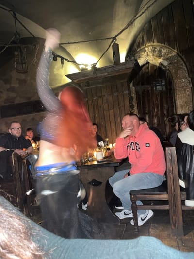 An indoor scene depicts a dimly lit, medieval-themed restaurant or pub in the Capital City of Prague, Czechia. A female performer with long, reddish-brown hair, wearing a shimmering blue-grey crop top exposing her midriff and a dark skirt with a blue waist sash, is captured in motion, actively dancing. Her hair and a raised arm exhibit significant motion blur, indicating movement. Several patrons are seated at heavy, dark wooden tables with carved bases and matching wooden chairs. To the left, a man with glasses and a beard, wearing a black shirt, watches the performance. To the right, a man with short hair and a beard, clad in a red hoodie and light blue jeans, sits with his chin resting on his hand, observing the dancer. Candles provide primary illumination on the tables, alongside ambient light from rustic fixtures. Glasses, mugs, and other tabletop items are visible. The environment features arched, aged plaster ceilings, rough textured walls, and dark wooden paneling. Chains, hooks, and suspended metallic elements resembling historical fixtures are visible overhead. A distinct wooden arched doorway is present on the right side of the frame.