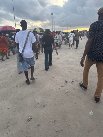 The image depicts a gathering of people on a sandy, outdoor area, likely in Satellite Town, Nigeria. The environment consists of a wide-open space with an overcast sky. Several people are visible, with one person in the foreground wearing a white shirt, denim shorts, and sandals. Another person in the upper frame is wearing a patterned shirt and brown pants. Several other people are in the background, with some appearing to be on motorcycles. There are streetlights visible. The sandy ground shows tire tracks and scattered debris.