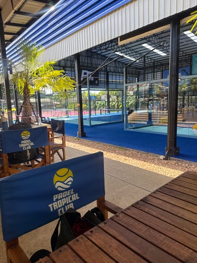 An outdoor seating area with several wooden chairs draped with blue fabric covers featuring a yellow and white logo that reads "PADEL TROPICAL CLUB". A wooden table is partially visible in the foreground. In the background, there are multiple indoor padel courts with blue and pink surfaces, enclosed by glass walls and metal structures. A palm tree with bright green fronds is situated to the left of the courts. Sunlight streams down from above, casting shadows. The scene appears to be at a sports club in Baan Bang Rak, Thailand.