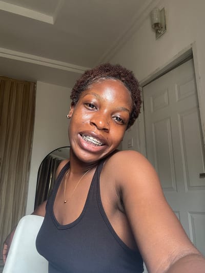 This indoor portrait features a young Black individual, likely female, in Lugbe, Nigeria. She has short, dark, curly hair and is smiling broadly, revealing braces on her upper teeth. A small stud piercing is visible on her left nostril, and she wears a small hoop earring in her left ear. Her skin has a dewy appearance, possibly due to lighting or natural oils.

She is wearing a black ribbed tank top with a subtle cutout design on the right strap, and a thin gold chain necklace with a small pendant rests on her chest. The background shows a light-colored ceiling with a recessed light fixture, and part of a white paneled door is visible. Below the door, there's horizontal wooden paneling, possibly furniture, and a white object (perhaps a chair or table) is partially seen in the lower right corner. The photo is taken from a slightly low angle, with the subject's head tilted, contributing to a casual and dynamic composition. The lighting is bright and appears even. The individual conveys a happy and confident emotion. No text is visible in the image.