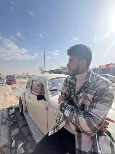 The image displays a young adult male posing outdoors on a sunny day in what is identified as Joubar Municipality, Syria. The man, positioned on the left side of the frame, has dark hair and a short beard. He is wearing a light-colored turtleneck sweater under an open green and white plaid flannel shirt and dark pants, with a silver ring visible on his right ring finger. He leans against a cream-colored vintage Volkswagen Beetle, with his arms folded across his chest, and is looking towards the right side of the frame.

The Volkswagen Beetle features chrome accents, black side mirrors, custom diamond-plate metal running boards, and a sticker resembling a cartoon skull with a hat on the lower part of the driver's side door. The car's interior, visible through the window, contains red/burgundy seating.

The foreground depicts a cracked concrete sidewalk strewn with litter and debris. In the background to the left, an older red pickup truck is parked on a dirt shoulder, and a series of distant buildings appear to be damaged or partially ruined. To the right, a commercial sign with Arabic text and logos, including Mercedes-Benz, is partially visible above a building structure. The sky is clear blue with scattered white clouds, and direct sunlight from the upper right creates a noticeable lens flare. A utility pole with overhead wires and a bare tree are also present in the middle ground.