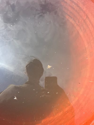 The image shows a reflection of a person holding a smartphone in murky water contained within an orange cylindrical object. The person's head and shoulders are visible as a silhouette, along with the distinct shape of the phone in their hand. Small white particles, possibly debris or light reflections, are scattered across the water's surface. The orange object appears to be a container, possibly a bucket or a planter, with ribbed texture visible on its interior surface towards the right side of the frame. The water's surface is disturbed, creating ripples and abstract patterns of light and shadow. The overall scene suggests an outdoor setting where the photographer has captured their reflection. The location in Berezovskii, Russia, is a detail that cannot be inferred from the visual information presented in the image.