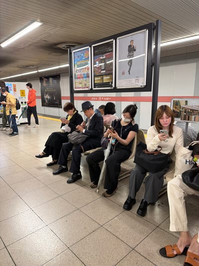 This image captures a subway or train station platform in Kyoto, Japan, identified by the "三条" (Sanjō) and "祇園四条" (Gion-Shijō) station names on a red and white sign below the advertisements, and "KEIHAN" branding on posters, indicating a Keihan Electric Railway line. The scene features seven individuals seated on a light-colored bench and two standing individuals on the left, all on a tiled platform. The ceiling is composed of light brown textured acoustic tiles with fluorescent strip lighting and some visible discoloration/stains. The walls feature a lower white section, a red horizontal stripe, and a darker gray upper section holding several illuminated advertising posters. From left to right, standing: 1. A man wearing a mustard yellow short-sleeved shirt over a green long-sleeved shirt, blue jeans, and black sneakers, with a dark backpack. He wears a white face mask and is looking down at his smartphone. 2. A man in an orange hoodie, dark pants, and light sneakers, standing further back and looking left. Behind them, a yellow and white vending machine and a multi-compartment white trash receptacle are visible. A dark green umbrella leans against the trash can. From left to right, seated on the bench: 1. A woman wearing a long black dress, black flat shoes, and a yellow scarf, intently looking at her smartphone. 2. An elderly man in a dark suit, white shirt, patterned tie, dark fedora-style hat, and black dress shoes. He holds a dark gray shoulder bag on his lap and is focused on his smartphone. 3. A woman wearing a black short-sleeved top, black pants, and a white face mask. She has a dark purple shoulder bag and a white folded umbrella leaning against her leg, and is looking at her smartphone. A colorful charm is attached to her phone. 4. A woman wearing a textured cream-colored long-sleeved top and dark gray pants. A black tote bag rests in front of her. She is engrossed in her smartphone. 5. Partially visible on the far right, a person wearing light cream-colored loose pants and brown leather sandals. A white canvas tote bag with "HA" visible is beside them, and a green packet (possibly containing tea or snacks) is on their lap. The floor is made of light beige square tiles with dark grout lines. All seated individuals, except the partially visible person on the far right, are actively engaged with their smartphones. No direct interaction