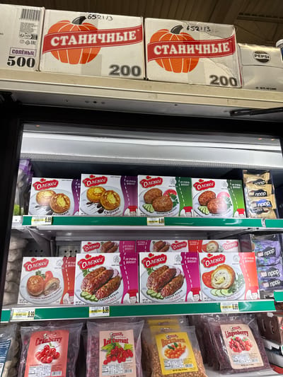 The image shows a refrigerated display case in a grocery store. The top shelf contains several boxes of food products, including "Станичные" (Stannichnye) brand products with images of pumpkins and the number "200" on the boxes, and Pepsi cans. The middle shelf displays boxes of "от ОЛЕЖКИ" (ot OLEZHKA) brand frozen food items, such as "СЫРНИКИ" (Syrniki - cheese fritters), "КОТЛЕТЫ МЯСНЫЕ" (Kotlety Myasnye - meat patties), "ЛЮЛЯ КЕБАБ" (Lyulya Kebab), and "КОТЛЕТЫ ПО-КИЕВСКИ" (Kotlety po-Kievski - Chicken Kiev). Some of these boxes have a price tag of $8.99. The bottom shelf contains bags of frozen berries, including "Cranberry," "Red Currant," "Bockthorn," and "Lingonberry." There are no people visible in the image. The overall environment suggests a retail setting, likely a supermarket or grocery store. Given the context of "Pepsi," it is possible this is a typical American grocery store. Specific location in Naperville, USA cannot be determined from the image alone, but it is a retail food environment.