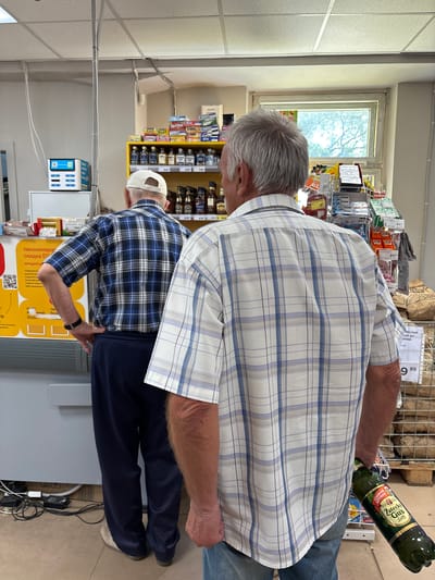 In a grocery store in Blagoveshchensk, Russia, two elderly men are browsing the aisles. The man in the foreground, with gray hair, is wearing a blue and white checkered shirt and holding a green bottle of "Zatecky Gus" beer. He is standing with his back to the camera. To his left, another man, wearing a blue and white checkered shirt and a white baseball cap, is also looking at the shelves. The store has bright fluorescent lighting and shelves stocked with various products, including alcohol, snacks, and household items. A window with trees visible outside is in the background. The floor appears to be tiled, and there are shopping carts and promotional displays throughout the store. The overall impression is of a typical shopping experience in a Russian supermarket.