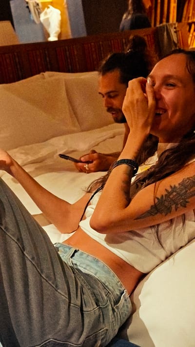 In Mù Cang Chải, Vietnam, an indoor scene depicts two individuals relaxing on a bed. In the foreground on the right, a woman with long dark hair, wearing a white sleeveless crop top and blue jeans, smiles broadly with her right hand covering her mouth. A dark, geometric tattoo is visible on her left upper arm, and a dark woven bracelet is on her left wrist. Her left arm rests on the white bedding. Behind her to the left, a man with dark hair tied in a bun and a beard looks down at a black smartphone held in his hands. He wears a light-colored top, and a ring is visible on his right ring finger. The bed is made with white sheets and pillows. In the background, a dark wooden headboard or wall paneling is present, and further back, blurred elements suggest additional furniture or items and soft, warm lighting.
