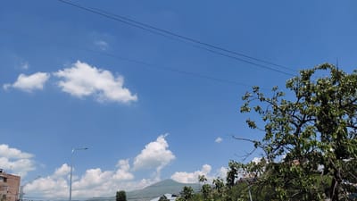 This media file captures a bright, sunny day in Abovyan, Armenia. The scene is dominated by a clear blue sky with scattered white cumulus clouds. Two dark power lines stretch across the upper portion of the image, adding a geometric element to the natural landscape. On the right side of the frame, a large tree with lush green leaves partially obscures the view, its branches reaching towards the sky. In the distance, a gently sloping mountain is visible, covered in a light green hue, suggesting it is a warm season.

Towards the lower left of the image, part of a multi-story building with a light brown facade is visible, hinting at an urban or semi-urban setting. A modern street lamp with a single light fixture stands tall, its metallic pole stark against the sky. Another, smaller street lamp is also visible further down the road. Below the mountain and the tree, the tops of other trees and some buildings can be seen, indicating a populated area.

The overall impression is one of a pleasant, ordinary day. The bright sunlight and clear sky suggest it is daytime, likely mid-morning to afternoon, and the weather is fair. There are no people or animals visible in the frame. The composition focuses on the expanse of the sky, the natural elements of the tree and mountain, and the subtle presence of human infrastructure.