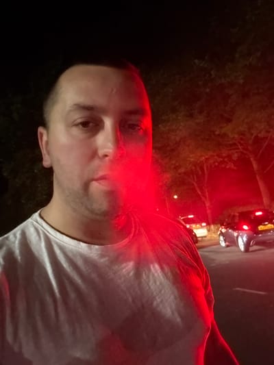 A man is seen in a selfie. The scene is dominated by a strong red lighting effect. The man has short hair, light skin, and stubble. He is wearing a light-colored t-shirt. In the background, there is a dark area of greenery and some blurred headlights of vehicles, suggesting a street at night. The location is the Borough of Spelthorne, United Kingdom.