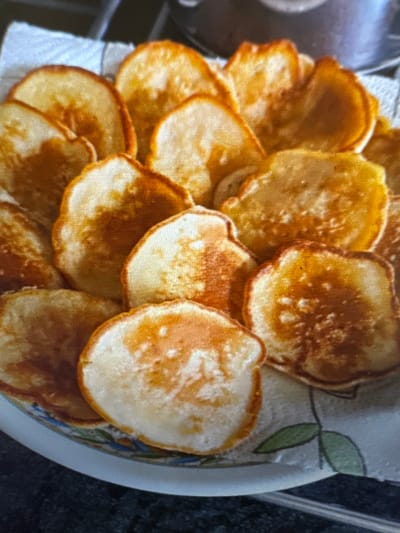 A plate of golden-brown pancakes is presented on a white paper towel, with a decorative ceramic plate peeking out from underneath. The pancakes are slightly irregular in shape, with some appearing thicker than others, and their surfaces show a varied texture with some lighter, slightly puffed-up areas and darker, crispier edges. The overall impression is of a home-cooked breakfast. The image provides no information about the city of Murcia, Spain.