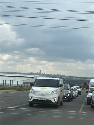 An overcast daytime scene captures a line of vehicles on an asphalt road in Midrand, South Africa, with the distant Johannesburg skyline visible on the horizon. The sky is predominantly grey with extensive cloud cover, traversed by multiple parallel overhead power lines.
In the immediate foreground, a white electric delivery van, bearing the South African license plate "LC 94 BZ GP" and a yellow triangular logo on its hood, is positioned slightly left of center, facing the viewer. Its integrated LED daytime running lights are illuminated. Directly behind this van, a dark grey SUV is visible, followed by a queue of several other vehicles, including cars and vans, extending into the mid-ground towards the right side of the frame. The front section of another white vehicle is partially visible on the far right edge.
To the left of the road, in the mid-ground, a series of industrial buildings with white roofs and grey walls are present, separated from the roadway by a strip of dry, sparse vegetation. To the right, a vegetated hillside is visible, along with a distant, partially obscured billboard. The background features a clear silhouette of the Johannesburg city skyline, including numerous buildings of varying heights and a distinct tall structure, consistent with the Hillbrow Tower. No individuals are discernibly visible; the scene depicts vehicles in a state of stationary or slow-moving traffic.