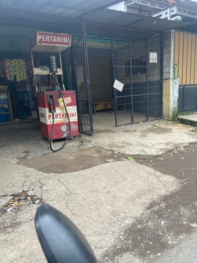 The image shows a gas station in Baleendah, Indonesia, located at the coordinates -7.021 latitude and 107.677 longitude. The gas station is a small, standalone structure with a red and white sign that says "Pertamini" in Indonesian.  A single gas pump is visible, and the station has a small, enclosed area with a gate. The pump is red and white with a sign that reads "Pasti Puas" (Guaranteed Satisfaction). The gas station is located on a narrow, paved street. There is a black motorcycle in the foreground, with its mirror visible in the bottom right corner of the image. The road surface is cracked and uneven. The area around the station is dusty and appears to be in an urban area. There are no people visible in the image.