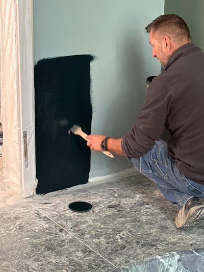 A man is painting a section of a light blue wall black. He is wearing a dark sweatshirt, blue jeans, and sneakers. A paintbrush is in his right hand. The floor is covered in a marble-like pattern and protected with plastic sheeting. A black circular object is visible in the floor near the wall. The location is unspecified, and there is no information indicating it is in Erba, Italy.