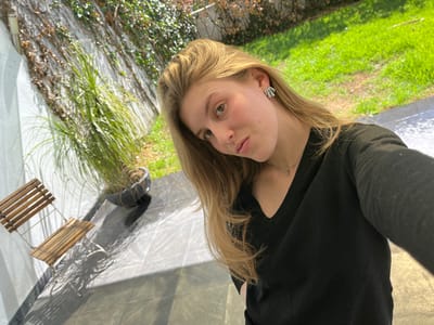 A young woman with long, blonde hair takes a selfie outdoors on a sunny day. She wears a black V-neck sweater and silver earrings. Behind her is a white wall covered in vines, a potted plant with long leaves, and a small wooden chair casting a shadow. A patch of green lawn is visible in the background. The ground appears to be concrete.