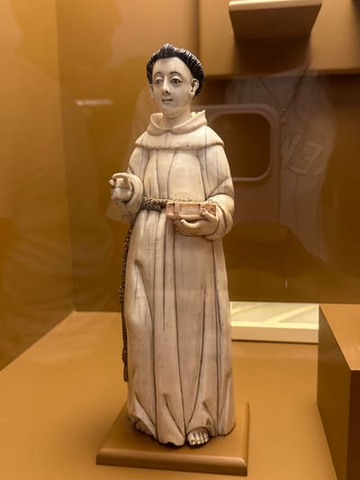 A finely carved statue of a male religious figure, likely a friar or monk, is presented in the image. The figure, possibly made of ivory, alabaster, or light-colored wood, is dressed in a flowing, cream-colored habit with detailed folds, secured at the waist by a dark, textured rope. His hair is dark and neatly styled, providing a strong contrast to his pale skin. The figure’s face conveys a serene and contemplative expression. He holds a small, rectangular book in his left hand, while his right hand is open with the palm facing upwards, possibly in a gesture of teaching or receiving.
The statue is displayed in an indoor setting, likely a museum or gallery, against a warm, ochre-colored background. Prominent reflections on a clear, protective surface (such as glass or acrylic) cover the foreground, showing abstract shapes and faint, indistinct letters like "EN" or "MEN" from the surrounding environment. The lighting is soft, creating subtle shadows that enhance the depth and detail of the sculpture. The overall scene suggests a historical or religious artifact, consistent with a display at a museum in Tepotzotlán, Mexico. There are no overt activities taking place, as it is a static display of an artwork.