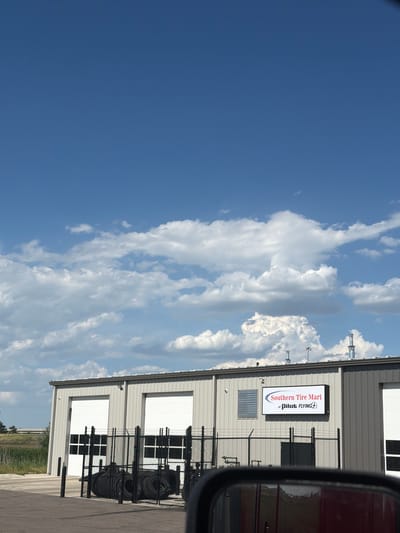 In North Platte, United States of America, a single-story, light gray industrial building with two large white roll-up garage doors is visible under a bright blue sky with scattered white clouds. A black chain-link fence encloses an outdoor area in front of the building, where several black tires are stacked. A sign on the building reads "Southern Tire Mart at Pilot Flying J." The image appears to be taken from the perspective of a vehicle, with the edge of a car's side mirror visible in the foreground. The environment is a flat, open area, with a grassy verge to the left of the building and some industrial structures or equipment visible on the roof.