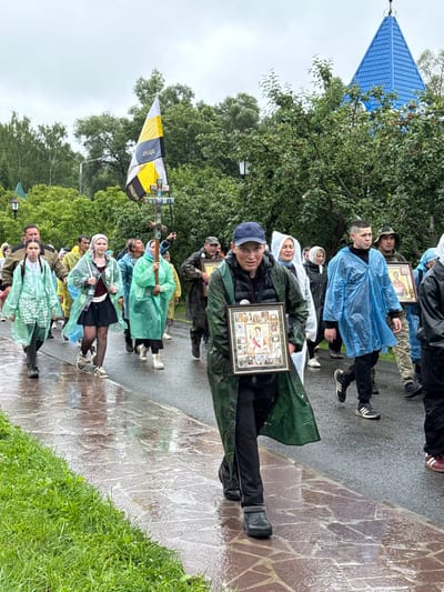 The image depicts a procession of people walking on a wet paved path, presumably in Местечко Раифа, Russia. The weather appears to be overcast and rainy, as many individuals are wearing transparent or colored plastic raincoats over their clothing. The people are of various ages and genders, and some are carrying religious icons. In the background, there are trees, a street lamp, and a building with a distinctive blue, conical roof. A flag with yellow and black diagonal stripes is visible towards the left side of the image. The ground is wet and reflective, indicating recent rainfall. The procession is moving forward, with individuals in focus walking towards the viewer.