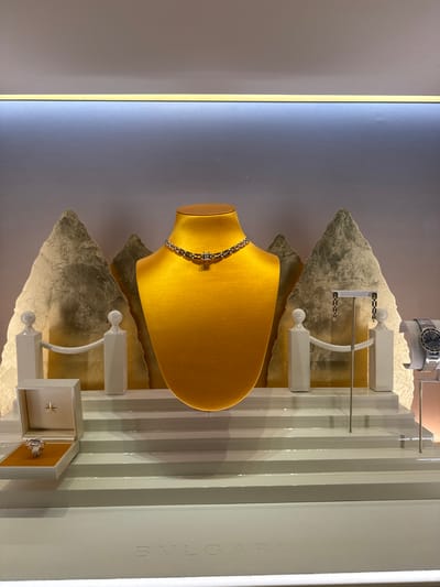 The image shows a high-end jewelry display inside a store. The display features a gold-colored mannequin bust wearing a silver necklace. To the left, a ring is presented in an open white box. To the right, a pair of earrings hangs from a stand, and a watch is displayed on a gray cushion. The backdrop consists of two abstract, textured golden shapes resembling mountains, with white decorative posts and railings. The display is set on multiple white tiered platforms. At the bottom of the image, the word "BVLGARI" is subtly embossed on the display's base. The lighting is soft and highlights the jewelry. The scene is set in a retail environment, likely a jewelry store, potentially located in the city of Nong Prue Subdistrict, Thailand, given the request. There are no people visible in the image.
