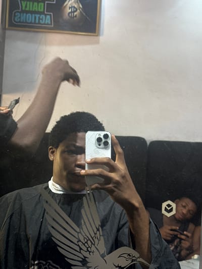 A mirror selfie captures a young Black male with short, dark, curly hair, holding a silver smartphone with a cracked screen protector. He is wearing a dark garment over a white high-neck top and has a neutral expression. The reflection shows him from the chest up, with his arm extending towards the phone.

The mirror itself appears slightly smudged and features a decorative, stylized wing or bird design etched onto its right side. On the left side of the reflection, a framed poster is visible, displaying a dollar sign symbol and reversed text that clearly reads "DAILY ACTIONS".

In the background of the reflection, another young Black male is seen lying down on what appears to be dark furniture, possibly a sofa or bed. He is looking at his own phone and has a faint smile. Near him, a white hexagonal symbol with an arrow-like design inside is visible on the dark surface.

The setting appears to be indoors, likely in a room in Ogwashi-Uku, Nigeria, under artificial lighting. The primary activity is the taking of a mirror selfie, while the secondary subject is relaxing.