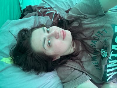 A young woman with dark hair and light green eyes lies on her back in a tent. She is wearing a grey t-shirt with green lettering that says "PREMIUM QUALITY 1998". Her head is tilted to her left, and she is looking towards the camera. The tent is made of green fabric. There are blankets and clothing items scattered around her. The image appears to have been taken indoors, possibly in a tent at a campsite. There are no visible signs of the location being in the city of Узюково, Russia.