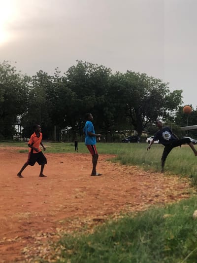 This vertically oriented image captures an outdoor scene in Nsugbe, Nigeria, featuring three males playing football on a dirt field.

In the background, one person, dressed in a dark t-shirt with a prominent white daisy graphic, is captured mid-air, performing an aerial kick on an orange-brown football. The ball is suspended above and to the left of their head, indicating a powerful strike. Lush green trees form a dense backdrop behind this active player, and a faint outline of a white vehicle is visible further back.

In the mid-ground, a second individual stands, wearing a blue t-shirt and dark shorts with red vertical stripes, seemingly observing the airborne player. Closer to the foreground, a third person is visible, wearing an orange t-shirt and dark shorts, also standing and facing the direction of the football action.

The playing surface is predominantly a reddish-brown dirt, characteristic of the region, flanked by sections of vibrant green grass and dense foliage. The sky above is bright and appears overcast or hazy, suggesting either an early evening glow or a cloudy day. The overall impression is one of casual, energetic outdoor activity. There is no discernible text present in the image.