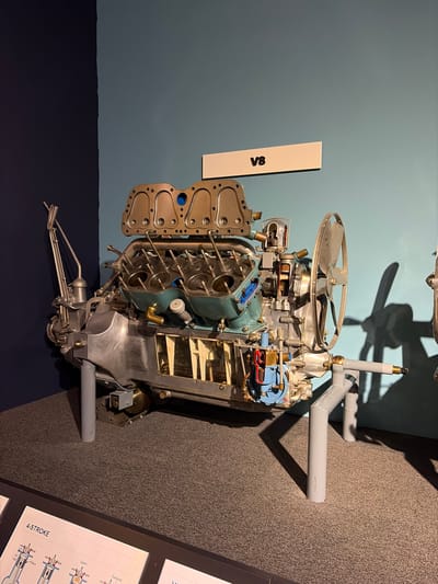 The image displays a detailed cutaway model of a V8 engine, presented as an exhibit in what appears to be a museum or educational setting in Wassenaar, Netherlands. The engine is positioned at an angle on a grey stand, showcasing its intricate internal mechanisms.
A large cooling fan with multiple blades is visible at the top of the engine. The engine block is partially opened and sectioned, revealing several cylinders with pistons and valve trains. Some internal components are highlighted with colors such as blue, red, and gold, likely to distinguish different parts or systems like fluid passages. A separate, unattached component resembling a valve cover with multiple bolt holes lies beside the main engine structure, further exposing the engine's internals. Pipes, possibly for exhaust or cooling, extend from the engine toward the dark grey carpeted floor.
The background consists of a plain light blue-grey wall, against which the engine and its stand cast shadows, suggesting indoor artificial lighting. To the left of the engine, a white rectangular sign with the black text "V8" is mounted on the wall. In the bottom right corner of the image, part of a black informational display panel is visible on the floor, featuring diagrams and the prominent text "4-STROKE," along with labels like "Exhaust," "Spark," "Piston," and "Air Fuel," indicating it explains the principles of a four-stroke engine cycle.
