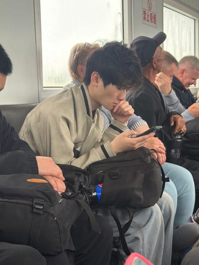 The image captures an interior scene, likely aboard a train, ferry, or bus, given the window and bench-style seating. Several individuals are visible, seated closely together in what appears to be a crowded compartment.

In the foreground, a young man with dark, curly hair is positioned on the left, wearing a light-colored jacket with dark stripes on the collar. He is engrossed in his smartphone, held in both hands, with one hand near his chin. Behind him, another person, with blonde hair and glasses, is partially visible, facing the window.

Further back, an older man wearing a dark baseball cap is seen, and behind him, another older man with gray hair is partially captured, seemingly engaged in animated conversation with an open mouth, wearing a light grey long-sleeved shirt. To the right of the young man, a black backpack with a visible Pepsi bottle is placed. Adjacent to this, hands with painted nails and a ring are holding a camera, suggesting photography or sightseeing. Towards the bottom right, another black backpack is visible, with a person's hands holding another person's hand next to it. Blue jeans are also visible on a seated person to the right.

The window to the left reveals an overcast, possibly rainy or foggy, exterior, with light streaks indicating precipitation or condensation on the glass. The general mood appears casual, with people engaged in their own activities or conversations.

A red and white sign on the wall near the window displays Chinese characters: "禁止吸烟" (Jìnzhǐ xīyān), meaning "No Smoking," along with the universal "no smoking" symbol. The presence of Chinese text in the context of Nizhneleninskoe, Russia, suggests a location near the Chinese border or a mode of transport catering to international travelers.