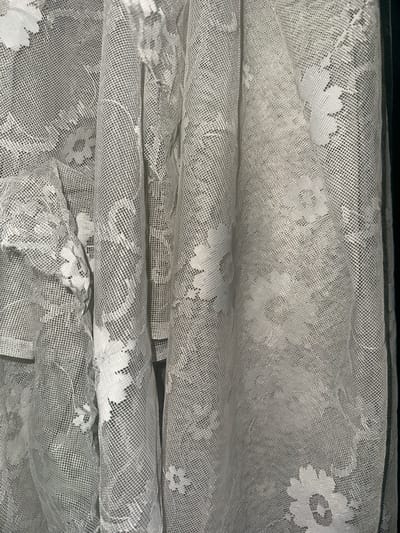 The image shows a close-up view of a white lace curtain. The curtain is draped, creating folds and shadows. The lace pattern features floral motifs, with small white flowers scattered across the fabric. The background is dark, creating a contrast with the light-colored lace. The scene is indoors, possibly in a home. There are no people or animals visible in the image. The photograph is taken from a slightly elevated angle, looking down at the draped fabric.