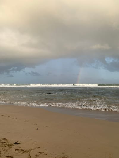 This image captures a serene beach scene in Kathaluwa, Sri Lanka. The foreground is dominated by a sandy beach with visible footprints, suggesting recent human activity. The middle ground features the ocean, with gentle waves rolling towards the shore, creating white foam at the water's edge. In the background, the sky is filled with dramatic, gray clouds, hinting at recent or impending rain. A faint rainbow arcs across the horizon, adding a touch of color to the overcast sky. The overall atmosphere is tranquil and somewhat moody, characteristic of a tropical coastal environment.