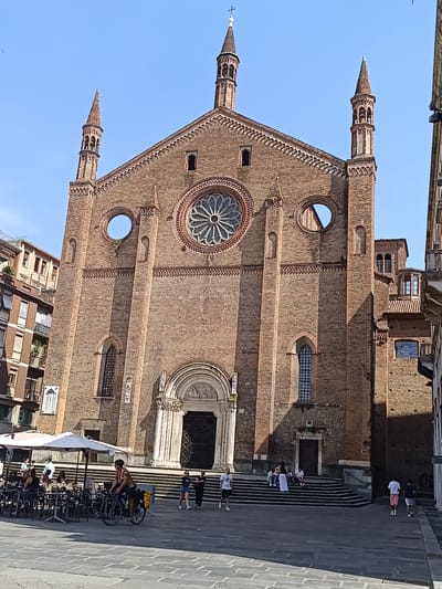 The image shows the Basilica of Sant'Antonino in Piacenza, Italy. The basilica, built of red brick, is viewed from its front facade. It has a central rose window and two narrower circular windows on either side. Twin towers flank the central peak of the roof. A grand entrance with a carved tympanum is at the base of the basilica. Wide steps lead up to the entrance.
In the foreground, a stone-paved square is populated with people. Some are sitting at tables under a white umbrella, others are walking across the square, and a cyclist with panniers rides by. Buildings adjacent to the basilica are visible on either side. The sky is clear and blue.