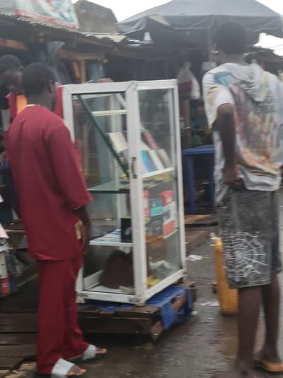 The image shows an outdoor scene in Madalla, Nigeria. A glass-fronted display case sits on a table in what appears to be a market area. Inside the case are packaged electronics, possibly mobile phones. Two men are visible. One man is wearing a red long-sleeved shirt and red pants. The other man is wearing a patterned t-shirt and shorts. The ground appears to be wet.