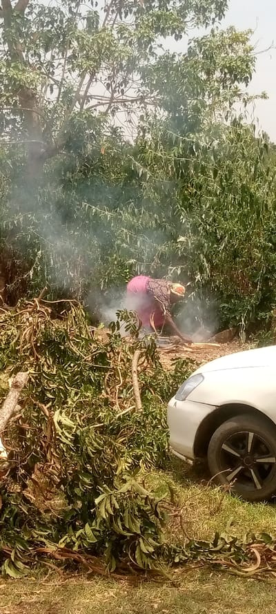 The image shows a woman wearing a pink and black patterned top, tending to a small fire in the woods. She is crouched down near the fire, which is producing smoke. There are branches and leaves scattered around the fire, and a white car is parked in the background. The location is in Burundu, Kenya, with coordinates 0.464 latitude and 34.74 longitude. The environment is lush and green, with dense vegetation.