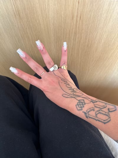 A person's hand is positioned on top of black fabric, likely clothing. The background consists of a light brown, wood-like surface. The hand has several rings: a gold ring on the thumb, a silver ring with a design on the index finger, and a thin gold band on the middle finger. The fingernails are long and squared, with a white design, some adorned with small gems. The person's arm is visible, featuring tattoos. One tattoo reads "Lucie" in script, and another shows the number "69" in a stylized font. The scene does not provide any information about the location in the city of Saint-Ouen-sur-Seine, France.