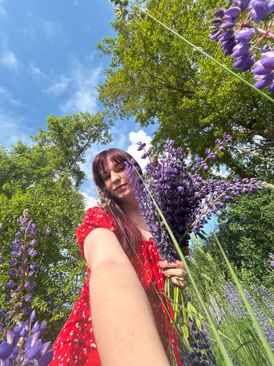 A young woman with long, auburn hair and bangs, wearing a red floral dress, is holding a bouquet of purple lupine flowers. She is outdoors, in a field of lupines, with lush green trees and a blue sky with scattered clouds visible in the background. The image is taken from a low angle, looking upwards. The woman appears to be taking a selfie, extending her arm towards the camera. The scene suggests a sunny day in a natural setting. The location is identified as the city of Garevaya, Russia.