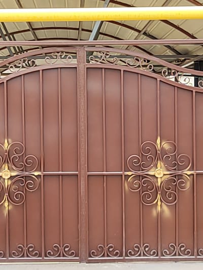 An outdoor medium shot captures a pair of tall, brown, wrought-iron double gates. The gates feature ornate scrollwork, with gold accents highlighting some of the decorative elements. The gates are closed. In the background, a yellow pipe runs horizontally above the gates, and a corrugated metal roof structure is visible. The scene appears to be in the city of Boraldai, Kazakhstan, suggesting a residential or commercial property with an enclosed yard or entrance.