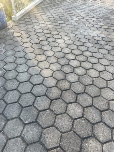 The image displays a textured surface of interlocking hexagonal concrete pavers. The pavers are grey and exhibit a slightly rough, granular finish. They are arranged in a repeating geometric pattern that extends across the frame, with some slight variations in shading due to lighting. In the upper left corner, a blue cylindrical object, possibly a trash can, is partially visible next to a white metal fence. Beyond the fence, green foliage and a bright, out-of-focus light source suggest an outdoor environment. The scene appears to be an outdoor walkway or patio area, likely in a public space given the presence of the fence and potential trash receptacle. The perspective is from a slightly elevated position looking down at the pavers, creating a sense of depth as the pattern recedes into the distance. The lighting suggests it is either morning or late afternoon, casting soft shadows. No people are present in the image. The location is identified as Bandungrejo, Indonesia, but no specific landmarks within Bandungrejo are visible.