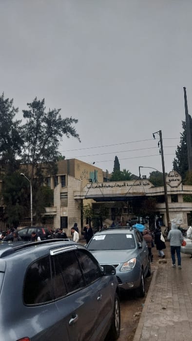 A cloudy outdoor scene shows a building with Arabic script, likely a university or institution, accompanied by trees and parked cars. Several people stand around near the vehicles, creating a crowded atmosphere on the street. The overall impression is of a gray and overcast day, with the architecture and people blending together under the gloomy sky.