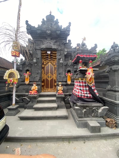 This image depicts an ornately decorated Balinese temple entrance, likely in Nusa Penida, Indonesia. The central focus is a grand, dark stone gateway with intricate carvings. A richly patterned cloth, predominantly red and white checkered with black accents, drapes over the entrance and is adorned with what appear to be offerings or decorative elements at the top. Several small, colorful statues of figures in traditional attire are placed on the steps leading up to the doorway, which itself is a dark wood with detailed golden embellishments.
To the left of the entrance, a tall, slender pole is decorated with dried palm fronds and a woven basket-like structure, also adorned with red and gold elements and possibly flowers. The architecture is typical of Balinese temples, with elaborate stone ornamentation and layered roofing structures visible above. The ground in front of the temple appears to be a paved or concrete surface, with some scattered debris. The sky above is bright and overcast, suggesting it is daytime, possibly morning or afternoon, with no strong shadows indicating a specific time. The overall atmosphere suggests a cultural or religious setting, likely prepared for a ceremony or festival, given the elaborate decorations. The image is taken from a slightly low angle, emphasizing the grandeur of the temple entrance. There is no visible text in the image.