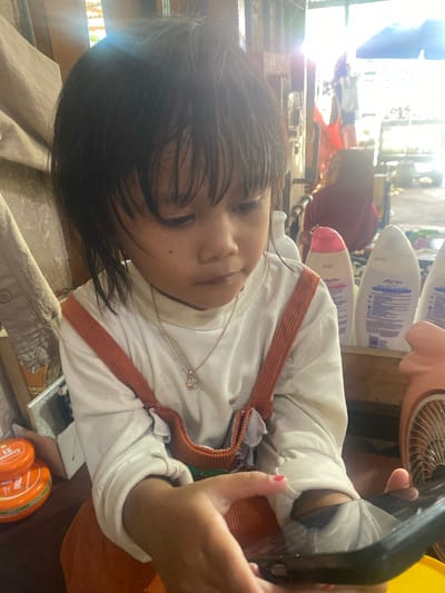 A young Indonesian child is lying down, with their head tilted towards the camera. They are wearing a white shirt with orange straps and a silver necklace. Their hair is dark and covers part of their face. The child is holding a black object, possibly a phone or tablet, in their hands. In the background, there are several bottles of lotion and a pink fan. Sunlight is streaming in from the left, creating a bright glare. The scene appears to be indoors, possibly in a home in Ta', Indonesia, given the context provided. The child's expression is neutral, and the overall mood of the image is casual and relaxed. The time of day appears to be daytime due to the strong sunlight.