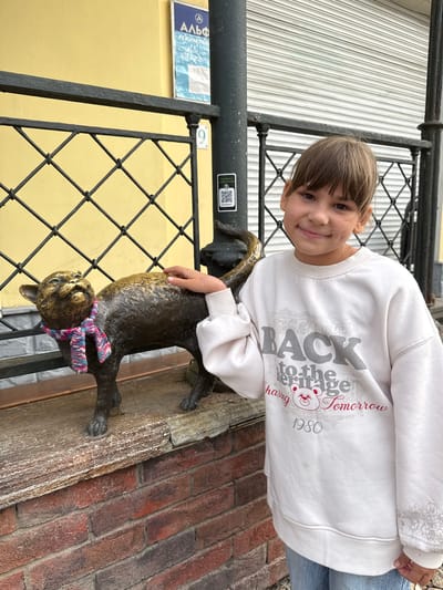 A young girl poses outdoors, smiling brightly directly at the camera. She has light brown hair with bangs and is wearing a white long-sleeved sweatshirt and blue jeans. Her right hand rests gently on the back of a bronze or dark metal cat statue, which features a colorful knitted scarf (pink, blue, and white stripes) tied around its neck.
The setting appears to be an urban or town environment. Behind the girl and the statue, there's a black metal fence with a diamond grid pattern. To the left, a light yellow building wall is visible, on which a blue and white sign with Cyrillic text is mounted. The sign prominently displays "Альфа" (Alfa) and a circled "3," with other smaller Cyrillic script below, consistent with the given location context of Заволжье, Russia. A small QR code sticker is also affixed to a black vertical pole near the fence. The ground or a lower wall surface to the right features distinct red brickwork. In the background, a white corrugated metal surface suggests a building facade or shutter.
The girl's sweatshirt features grey and red text: "BACK to the... (obscured word, possibly 'Future' or 'Forge') chasing tomorrow 1980," along with a cartoon bear head graphic. The time of day appears to be daytime, likely overcast given the soft lighting. The overall mood is cheerful, with the girl's happy expression and casual interaction with the public art.
