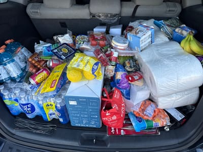 The image shows the trunk of a car filled with groceries. The car appears to be parked in a residential area, possibly a driveway or street in Prospect Heights, Illinois. The groceries include a variety of items such as bottled water, beverages, produce like bananas and blueberries, packaged meats, dairy products, and canned goods. There are also some non-food items such as a cleaning brush set and a large roll of plastic wrap. The overall scene suggests a recent grocery shopping trip.
