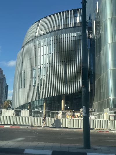 This media file captures a sunlit street scene in Givatayim, Israel. The foreground features a dark lamppost. Behind it, a modern, curved building with a facade of horizontal metallic louvers dominates the view. The building's reflective surface mirrors the sky and surrounding structures.

To the right of the building, a street is visible, bordered by a sidewalk. A construction site is present, indicated by temporary fencing and corrugated metal panels. In the middle ground, a person is walking along the sidewalk.

In the background, the sky is a clear, bright blue, suggesting a daytime setting. The presence of the construction site and the clear weather indicate ongoing development and a pleasant day. The architectural style of the main building suggests a contemporary urban environment.

Visible text includes Hebrew lettering on signs within the construction area. One sign, partially obscured, appears to read "הדר". Another sign, clearer, also has Hebrew text, likely related to the construction project. The overall impression is one of an active, developing urban area under a bright sky.