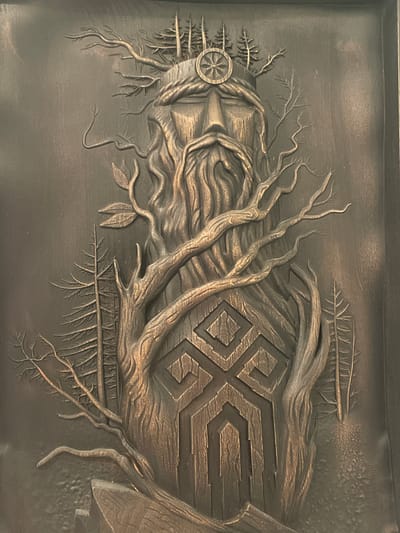 The image depicts a close-up, full-frame view of a bas-relief sculpture. The sculpture features a stylized, anthropomorphic figure with a long, flowing beard made of wood grain textures. The figure wears a crown composed of tree branches and small evergreen trees. A circular symbol with radiating lines, resembling a sun or wheel, is embedded in the center of the crown. The figure's body is intertwined with thick, gnarled branches and smaller, delicate twigs. On the chest of the figure, a geometric symbol with a diamond shape in the center and layered polygonal patterns is carved. The overall color palette is monochromatic, with variations in light and shadow creating a sense of depth and texture, suggesting a metallic material such as bronze. The background is a plain, textured surface that supports the relief. The sculpture is lit from the front, casting shadows that highlight the intricate details of the carving. There are no people, actions, or interactions visible in the image. The sculpture appears to be located in the city of Pushkin, Russia, likely within a museum or an art installation, although the specific location within Pushkin is not indicated by the image itself.