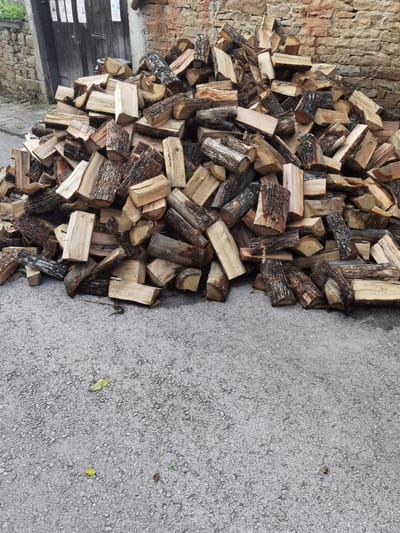 A large pile of chopped firewood is stacked on a grey asphalt surface. The wood is of various sizes and types, with some pieces showing rough bark and others with smooth, cut ends. In the background, a rough stone wall made of beige bricks is visible, and behind that, a weathered wooden door. The environment appears to be outdoors, likely in a rural or semi-rural setting, consistent with the village of Lomets, Bulgaria. There are no people or animals in the image. The lighting suggests it is daytime, with some shadows cast by the wood.