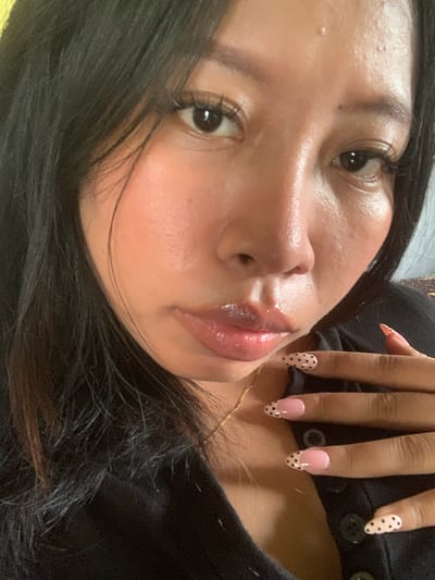 A close-up, selfie-style shot shows a young woman with dark hair, a fair complexion, and dark eyes. She is wearing a black top and has long, sculpted fingernails with a pink and black polka dot design. The woman's face is slightly turned, and her lips are glossy. She is resting her hand on her chest, and a gold necklace is visible. The background appears to be a neutral-toned fabric. The overall impression is of a casual, self-portait moment.